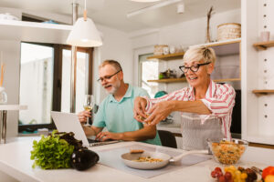 Mature couple in kitchen, man on laptop, wife cooking, making retirement plans